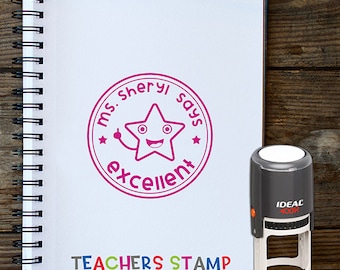 May include: A pink teacher's stamp with a cartoon star saying "Ms. Sheryl says excellent". The stamp is on a white notebook with the words "TEACHERS STAMP" printed below it. The stamp is next to a grey self-inking stamp.