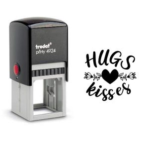 Peut inclure: Un tampon encreur noir et gris avec le texte "HUGS kisses" dans une police décorative, avec un cœur et des motifs floraux. Le tampon a un bouton rouge et la marque "trodat printy 4924".