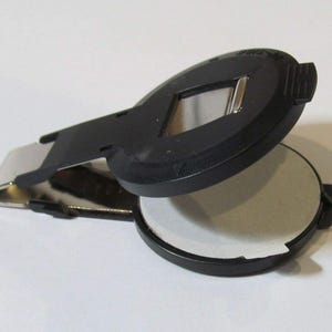 May include: A black and silver ink pad with a hinged lid. The lid has a rectangular opening for applying ink to a stamp.