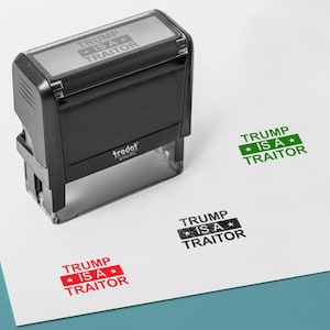 May include: A black Trodat stamp with the text "TRUMP IS A TRAITOR" printed on the top. The stamp has made impressions in red, green, and black ink on a white surface. The stamp is a political statement.