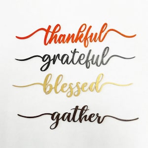 May include: Four wooden signs with the words "thankful", "grateful", "blessed", and "gather" written in cursive. The signs are painted in different colors: orange, black, gold, and brown.