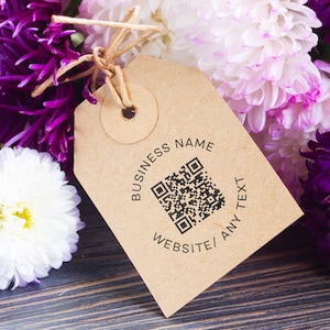 Puede incluir: Una etiqueta de papel marrón con un código QR y las palabras "BUSINESS NAME" y "WEBSITE/ ANY TEXT" impresas en negro. La etiqueta está atada con hilo y colocada sobre un fondo de flores moradas, blancas y rosas.