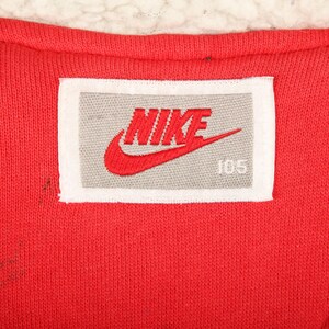 Pu&ograve; includere: Primo piano di un tessuto rosso con un'etichetta bianca e grigia. L'etichetta presenta il logo Nike in rosso e la parola "NIKE" in rosso, insieme al numero "105". Il tessuto ha un aspetto strutturato.