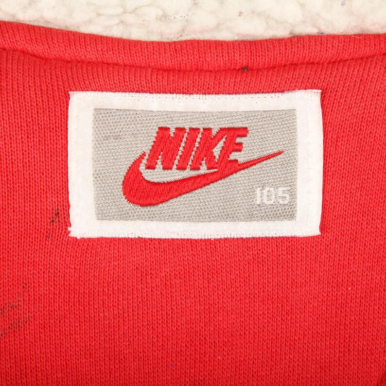 Pu&ograve; includere: Primo piano di un tessuto rosso con un'etichetta bianca e grigia. L'etichetta presenta il logo Nike in rosso e la parola "NIKE" in rosso, insieme al numero "105". Il tessuto ha un aspetto strutturato.