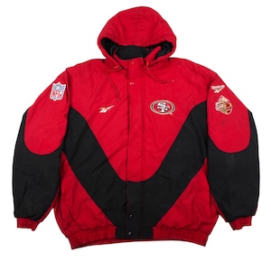 Może przedstawiać: Czerwono-czarna kurtka z kapturem z logo San Francisco 49ers na piersi, naszywką NFL na prawym rękawie, logo Reebok na piersi i lewym rękawie oraz pamiątkową naszywką z okazji 50-lecia umieszczoną na lewym rękawie kurtki.