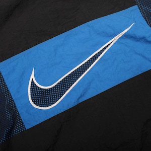 May include: Black and blue athletic jacket with a large white-outlined Nike swoosh logo. The logo is centered on a diagonal blue panel, with black panels on either side. The jacket has a mesh detail.