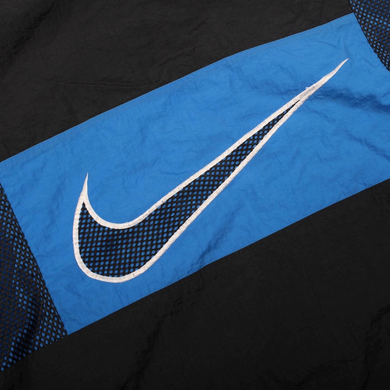 May include: Black and blue athletic jacket with a large white-outlined Nike swoosh logo. The logo is centered on a diagonal blue panel, with black panels on either side. The jacket has a mesh detail.