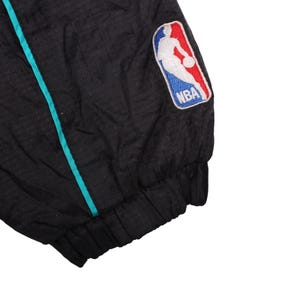 May include: Black jacket with a teal stripe and the NBA logo patch. The jacket has a textured pattern and a ribbed cuff. The NBA logo features a basketball player in white, with red and blue accents.