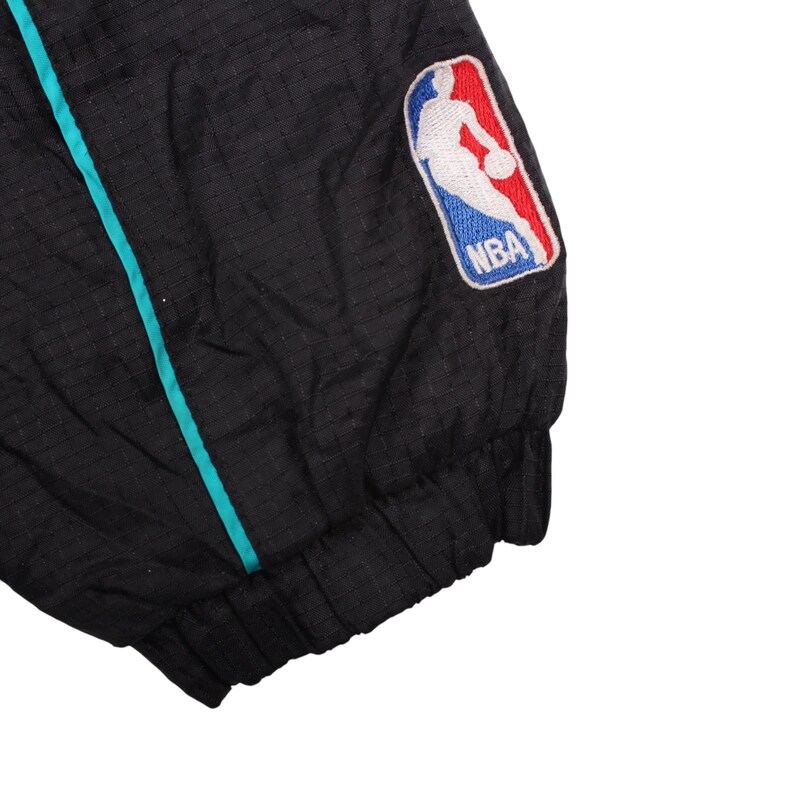 May include: Black jacket with a teal stripe and the NBA logo patch. The jacket has a textured pattern and a ribbed cuff. The NBA logo features a basketball player in white, with red and blue accents.