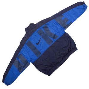May include: A long-sleeve windbreaker jacket in navy blue and royal blue. The back features the word NIKE in large, dark navy block letters across the shoulders and sleeves, with a small navy swoosh logo positioned between the letters.