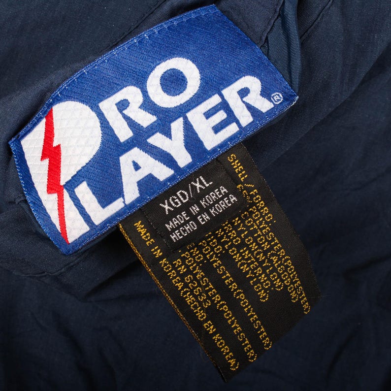 Op de afbeelding: Close-up van een marineblauw kledinglabel met de witte tekst "PRO PLAYER" en een rood bliksemontwerp. Het label bevat ook informatie over de maat en de materiaalsamenstelling.