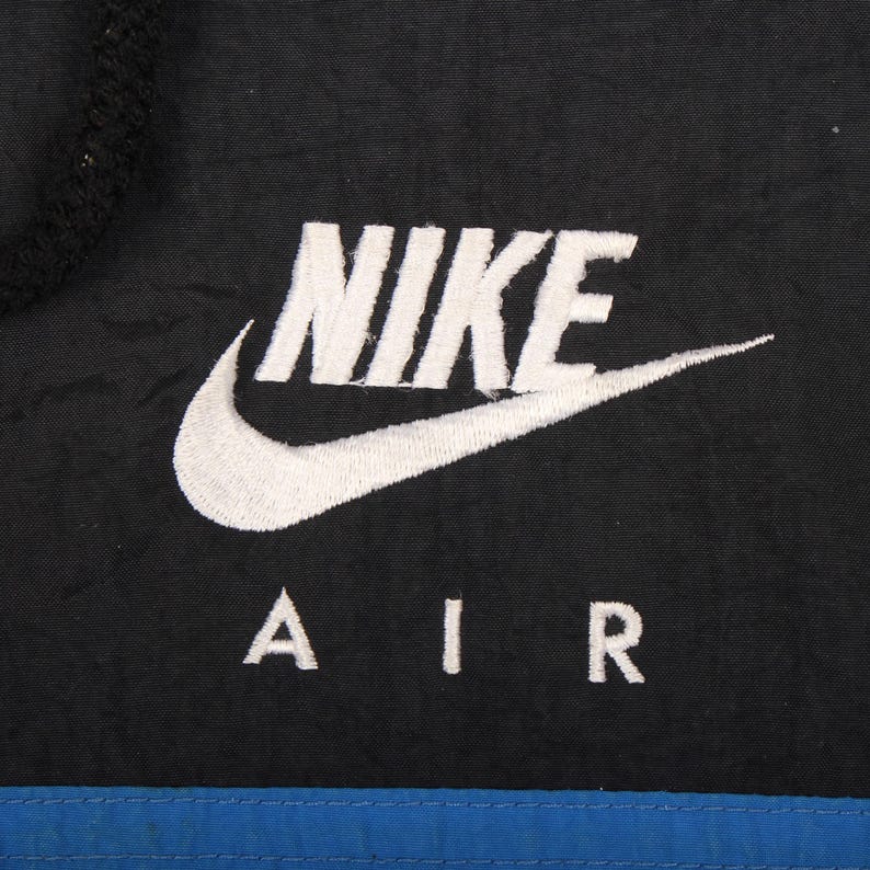 May include: Close-up of a black jacket with the white Nike logo and the word "NIKE" embroidered on the front. Below the logo, the word "AIR" is also embroidered in white. A blue stripe is at the bottom.