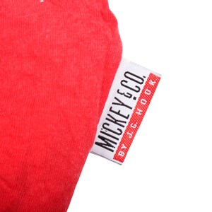 May include: A bright red fabric garment featuring a white rectangular clothing tag. The tag displays the black text MICKEY & CO. followed by the smaller text BY J.G. HOOK, with a red border on the right side of the tag.