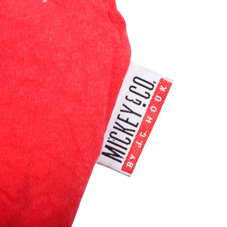 May include: A bright red fabric garment featuring a white rectangular clothing tag. The tag displays the black text MICKEY & CO. followed by the smaller text BY J.G. HOOK, with a red border on the right side of the tag.