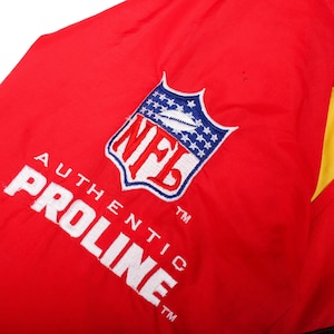 May include: A bright red jacket featuring an embroidered NFL shield logo in red, white, and blue, positioned above the white embroidered text AUTHENTIC PROLINE. The fabric has a smooth texture with a visible yellow panel on the right side.