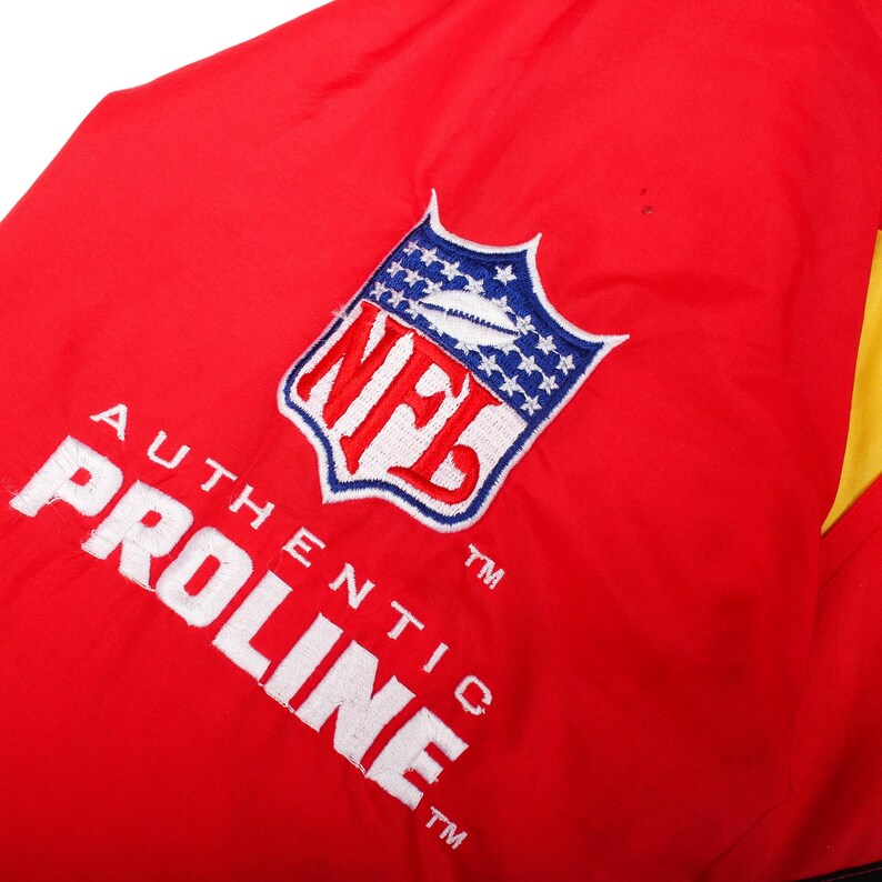 May include: A bright red jacket featuring an embroidered NFL shield logo in red, white, and blue, positioned above the white embroidered text AUTHENTIC PROLINE. The fabric has a smooth texture with a visible yellow panel on the right side.