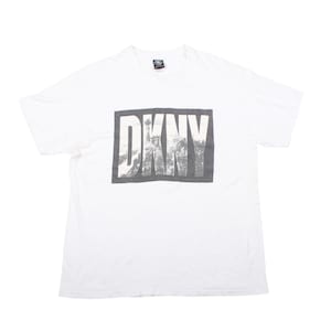 May include: White t-shirt with a large rectangular graphic displaying the letters "DKNY" in white. The letters are set against a grey background with a blurred cityscape design. The shirt has a crew neck and short sleeves.