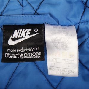 May include: Close-up of a blue quilted fabric with black stitching. Two tags are visible: a black Nike tag with white text and a white tag with care instructions and size information. The Nike tag reads "made exclusively for FOOTACTION".