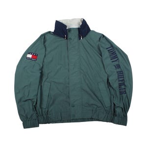 以下が含まれることがあります： 白とネイビーの襟が付いた、ティール色のトミーヒルフィガーのジャケット。左胸にトミーヒルフィガーのロゴ、左袖にブランド名がプリントされています。ボタン留めフロントと2つのフロントポケットが特徴です。