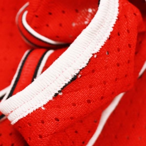 May include: Close-up of red fabric with a white trim and black stripes. The fabric has a mesh pattern with small holes.