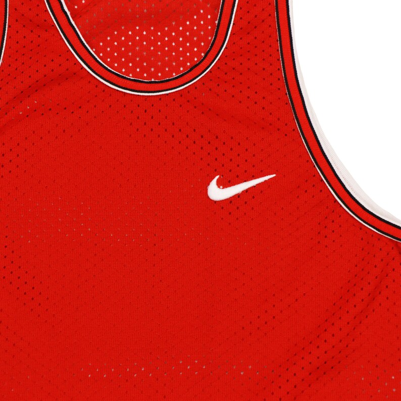 May include: Red basketball jersey with a white Nike swoosh logo. The jersey is made of a mesh material and has a black and white trim around the neck.