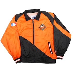 May include: A zip-up windbreaker jacket in bright orange and black with long sleeves, a collar, and ribbed cuffs. The front features an embroidered Baltimore Orioles logo, while the right sleeve displays a Major League Baseball logo patch.