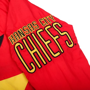 May include: A bright red jacket sleeve featuring the text Kansas City CHIEFS embroidered in bold yellow lettering with black outlines. The fabric appears to be a smooth, lightweight material with a yellow panel visible at the bottom edge.