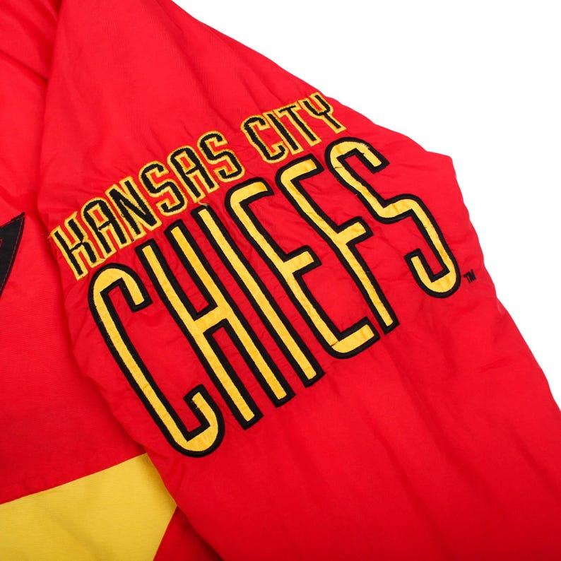 May include: A bright red jacket sleeve featuring the text Kansas City CHIEFS embroidered in bold yellow lettering with black outlines. The fabric appears to be a smooth, lightweight material with a yellow panel visible at the bottom edge.