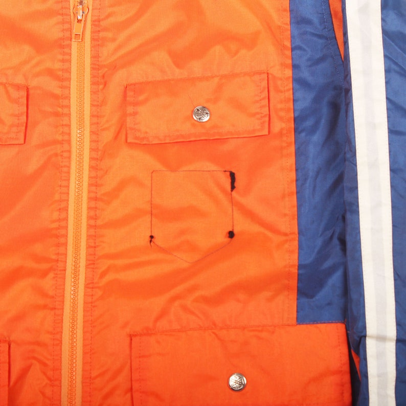 K&ouml;nnte beinhalten: Eine leuchtend orangefarbene Jacke mit einem blauen und wei&szlig;en Streifen an der Seite. Die Jacke hat einen Rei&szlig;verschluss und mehrere Taschen.