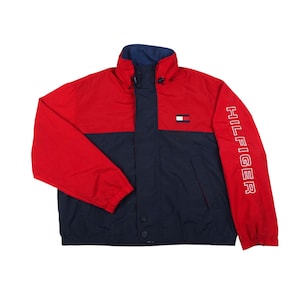 Op de afbeelding: Een rode en marineblauwe jas met het woord "HILFIGER" verticaal in het wit gedrukt op de rechter mouw. De jas heeft een hoge kraag en een klein logo op de borst.