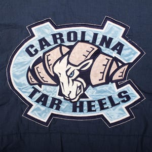 Op de afbeelding: Een marineblauwe patch met de woorden "CAROLINA TAR HEELS" in witte letters. De patch heeft een lichtblauwe rand en een centraal beeld van een ramshoofd in wit, beige en marineblauw.