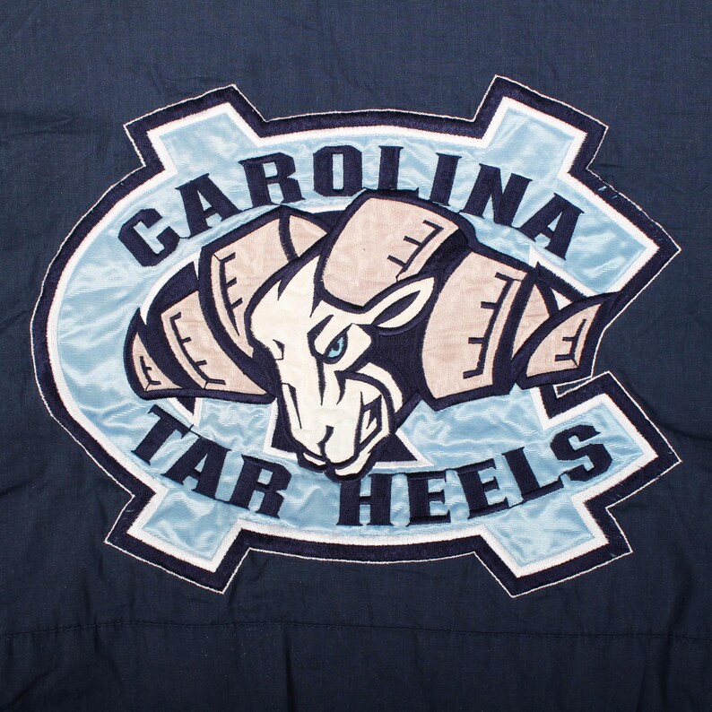 Op de afbeelding: Een marineblauwe patch met de woorden "CAROLINA TAR HEELS" in witte letters. De patch heeft een lichtblauwe rand en een centraal beeld van een ramshoofd in wit, beige en marineblauw.