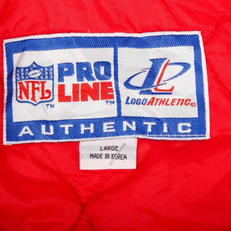 May include: A red quilted garment interior featuring a white rectangular tag with blue borders. The tag displays the NFL Pro Line logo, the Logo Athletic logo, and the word AUTHENTIC. A smaller white tag below indicates size LARGE and MADE IN KOREA.