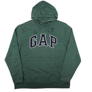 Könnte beinhalten: Ein waldgrüner Kapuzenpullover mit langen Ärmeln, der auf der Brust das Wort GAP in großen, dunkelmarineblauen Blockbuchstaben mit weißer Umrandung zeigt. Das Kleidungsstück hat eine Kängurutasche vorne und passende grüne Kordelzüge.