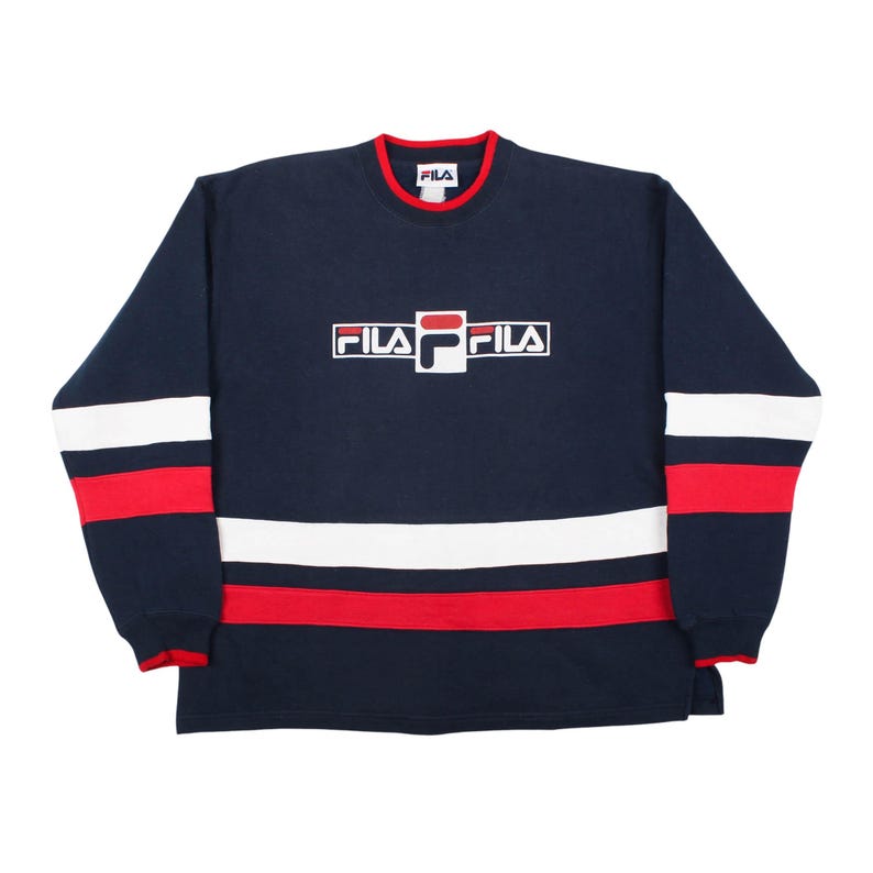 vintage 90s navy blue Fila crewneck sweatshirt on a white background, featuring a bold white and red embroidered Fila logo in the center chest and matching solid horizontal white and red stripes across the lower body and sleeves with a red ringer collar