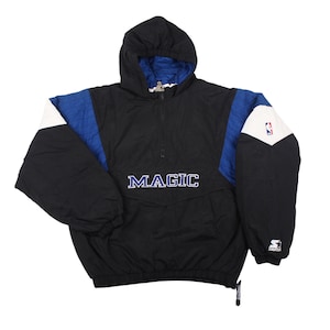 Op de afbeelding: Zwarte, blauwe en witte anorakjas met capuchon met het woord "MAGIC" over de borst. De jas heeft een halve ritssluiting, een blauw en wit kleurblokontwerp op de mouwen en het NBA-logo op de rechter mouw.