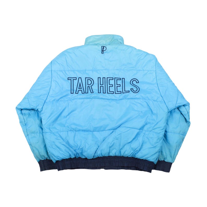 Op de afbeelding: Lichtblauwe pufferjas met de tekst "TAR HEELS" in marineblauw op de rug. De jas heeft een marineblauwe kraag en manchetten. Een klein "P"-logo is zichtbaar op de bovenrug.