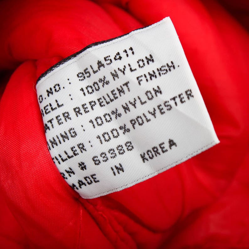 May include: A white rectangular garment care label sewn onto a bright red nylon fabric. The text reads: O.NO.: 95LA5411, SHELL: 100% NYLON, WATER REPELLENT FINISH, LINING: 100% NYLON, FILLER: 100% POLYESTER, RN # 63388, MADE IN KOREA.