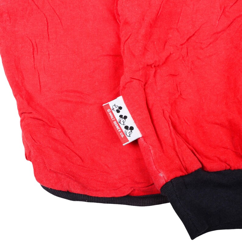 May include: A bright red garment with a black ribbed cuff at the bottom edge. A small white rectangular fabric tag is attached to the side, featuring three black Mickey Mouse head silhouettes and the text WALT DISNEY CO. printed in black.