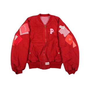 May include: A red bomber jacket with a zipper closure and a gingham lining. The jacket features patches with text and a large pink "P" on the chest. The sleeves have a puffy design, and the jacket has a ribbed hem and cuffs.