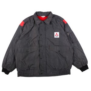 May include: A charcoal gray quilted jacket featuring a red interior lining, a fold-down collar, and black ribbed cuffs. The front displays a white Mitsubishi Motors logo patch, two large flap pockets, a snap-button closure, and red shoulder accents.