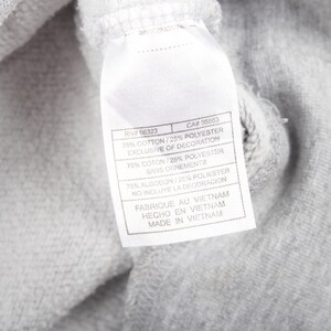 May include: A close-up shot of grey fabric with a white label attached. The label shows text including "75% COTTON/25% POLYESTER" and "MADE IN VIETNAM". The fabric is likely part of a piece of clothing.