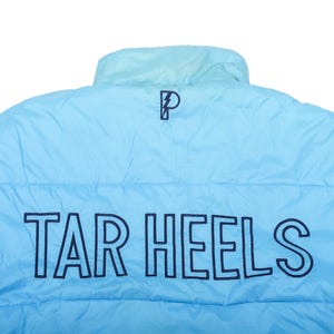 Op de afbeelding: Lichtblauwe pufferjas met de woorden "TAR HEELS" in marineblauwe blokletters. Een marineblauw "P"-logo staat boven de tekst. De jas heeft een opstaande kraag.