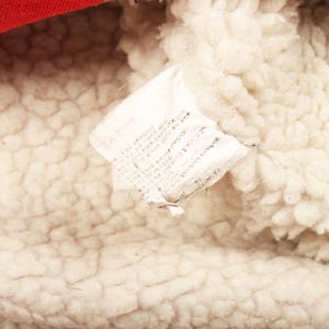 Pu&ograve; includere: Primo piano di un'etichetta di tessuto bianco con testo, attaccata a un materiale color crema, strutturato e soffice. Una sezione di tessuto rosso &egrave; visibile nella parte superiore. L'etichetta sembra essere un'etichetta di cura.