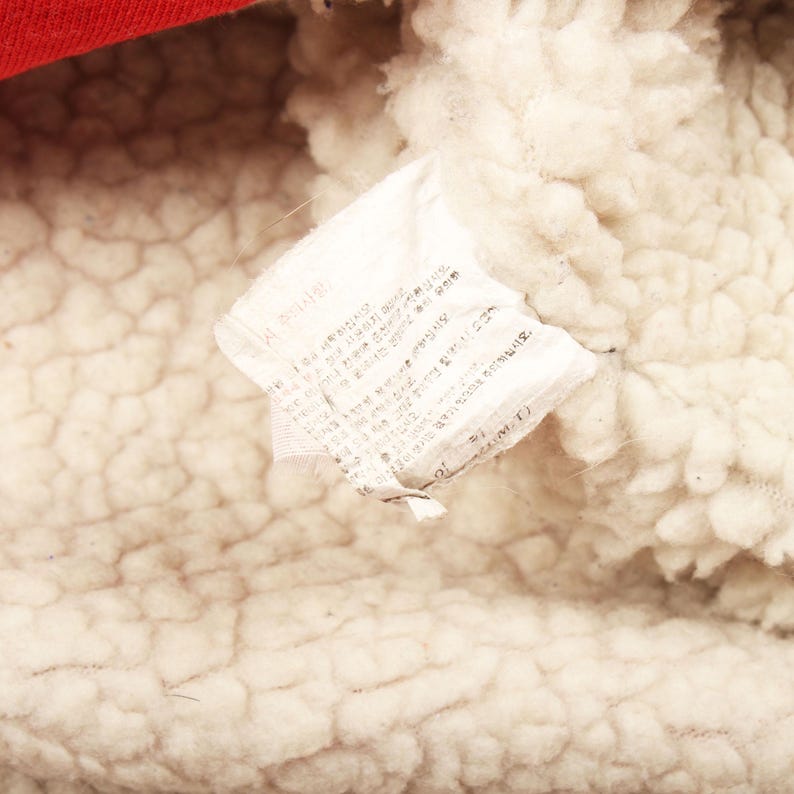 Pu&ograve; includere: Primo piano di un'etichetta di tessuto bianco con testo, attaccata a un materiale color crema, strutturato e soffice. Una sezione di tessuto rosso &egrave; visibile nella parte superiore. L'etichetta sembra essere un'etichetta di cura.