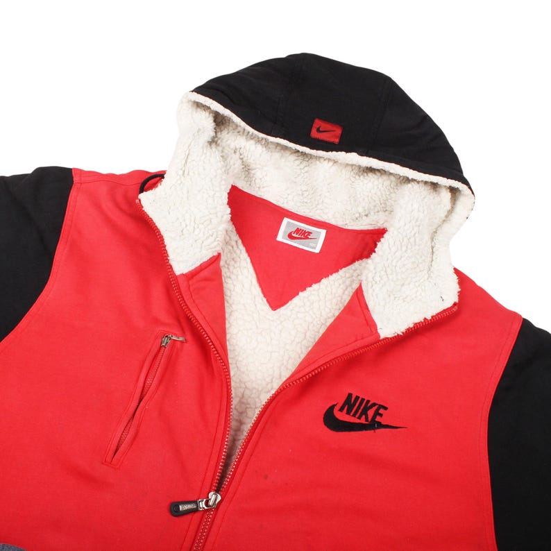 Pu&ograve; includere: Giacca con cappuccio Nike rossa e nera con fodera in pile bianco. La giacca presenta una cerniera completa, una piccola tasca con cerniera e il logo Nike nero sul petto. Il cappuccio &egrave; nero con un piccolo logo Nike rosso.