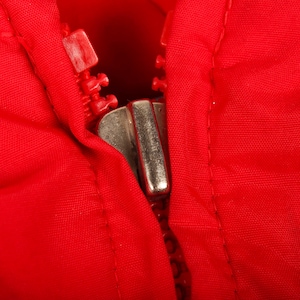 May include: A bright red fabric garment featuring a metallic silver zipper pull and matching red plastic zipper teeth. The zipper is partially closed, showing the interlocking mechanism against the vibrant red textile material of the clothing.