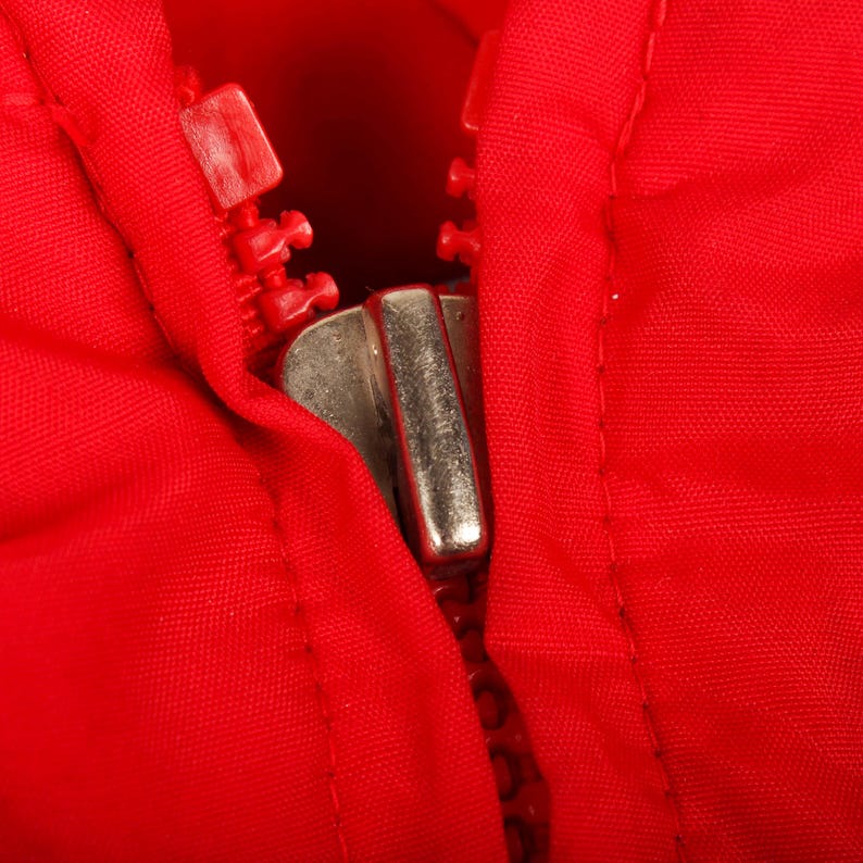 May include: A bright red fabric garment featuring a metallic silver zipper pull and matching red plastic zipper teeth. The zipper is partially closed, showing the interlocking mechanism against the vibrant red textile material of the clothing.
