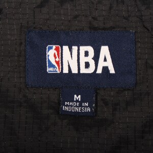 May include: A close-up shot of a navy blue label featuring the NBA logo and text. Beneath it, a smaller navy label displays the letter "M" and the text "MADE IN INDONESIA". The background is black.
