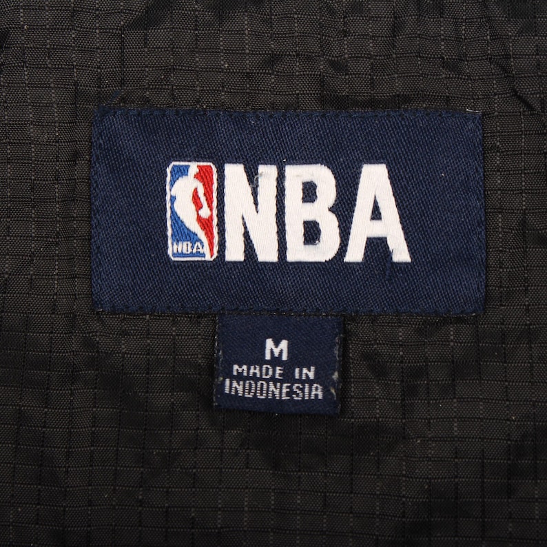 May include: A close-up shot of a navy blue label featuring the NBA logo and text. Beneath it, a smaller navy label displays the letter "M" and the text "MADE IN INDONESIA". The background is black.